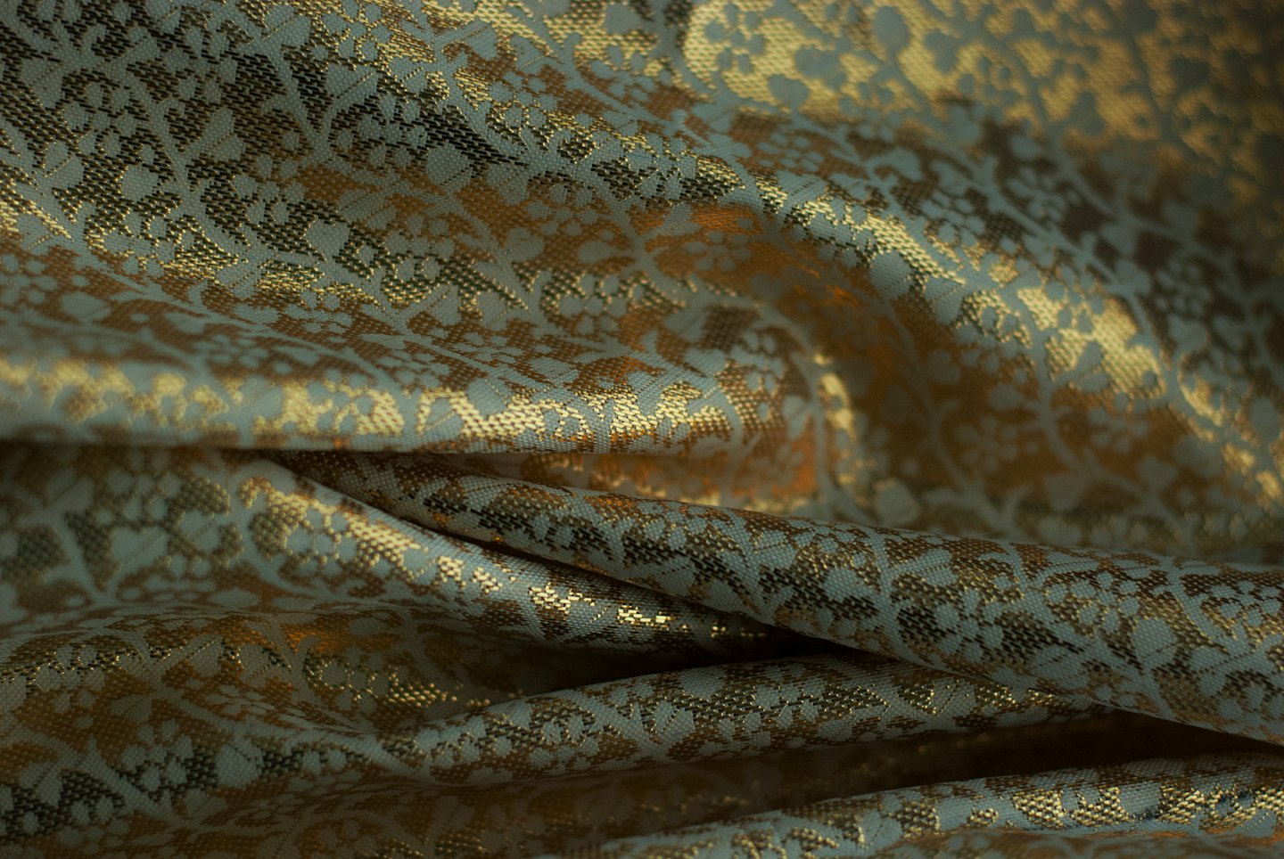 Intricato motivo floreale dorato su tessuto drappeggiato grigio-verdastro.